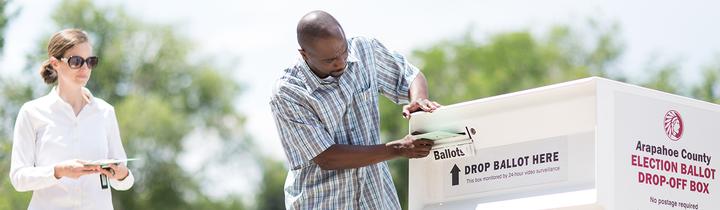 Male worker inserts ballot into outside ballot box while female approaches behind with ballot envelope in hand