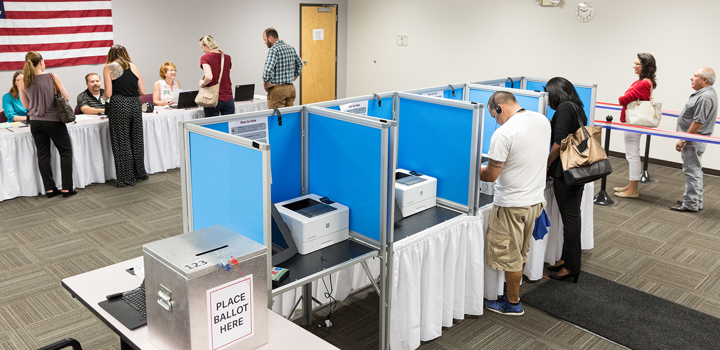 Four voters stand at check-in table, two voters stand at blue voting booths and two voters stand in line