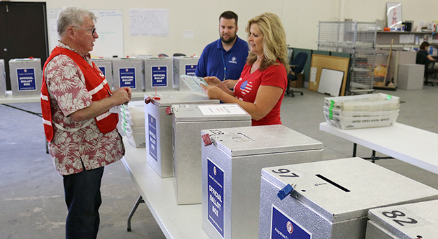 Three election workers stand next to table with silver ballot boxes