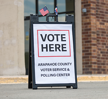 A sign stands outside a brick building with the words Vote Here. Arapahoe County Voter Service and Polling Center.