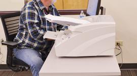 An election worker sits in front of a computer screen and a large scanner stacked with ballots