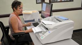 An election worker sits in front of a computer screen and a large scanner stacked with ballots