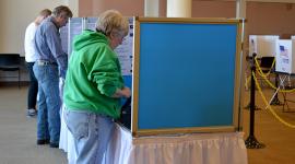 Three voters stand at blue voting booths and cast votes on electronic tablets