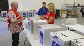 Three election workers stand next to table with silver ballot boxes