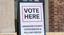 A sign stands outside a brick building with the words Vote Here. Arapahoe County Voter Service and Polling Center.