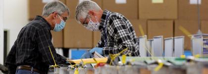 A pair of two men in face masks looking at retrieving ballots for a random audit from a row of ballot boxes.